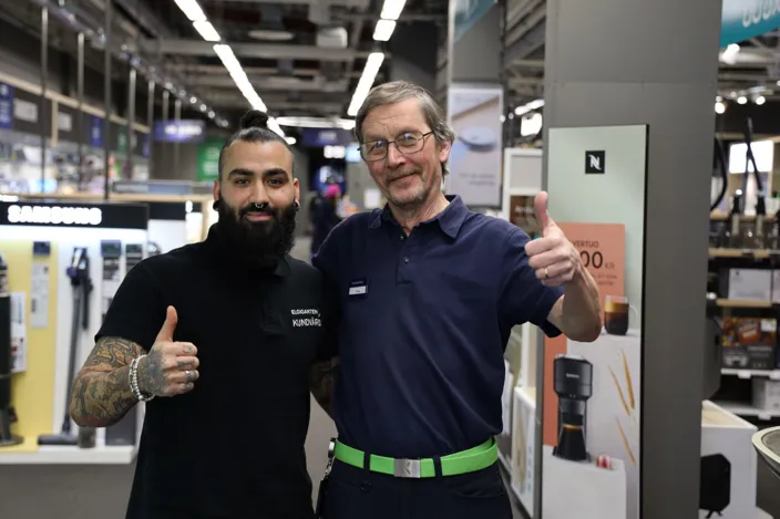 Bild på två män som står i en elgigantenbutik. De håller om varandra med en arm, den andra visar tummen upp. Båda tittar in i kameran och ler.