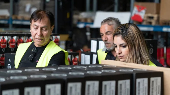 Två samhall-medarbetare och Nooshi står bakom några produkter. Man ser deras huvuden sticka upp bakom lådorna som täcker deras kroppar. De bär reflexvästar. 