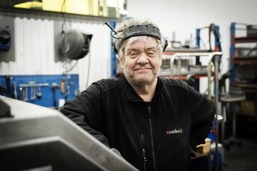 En medarbetare står i en verkstadsmiljö med skyddsutrustning på huvudet. I bakgrunden syns verktyg, arbetsstationer och industrimaskiner.