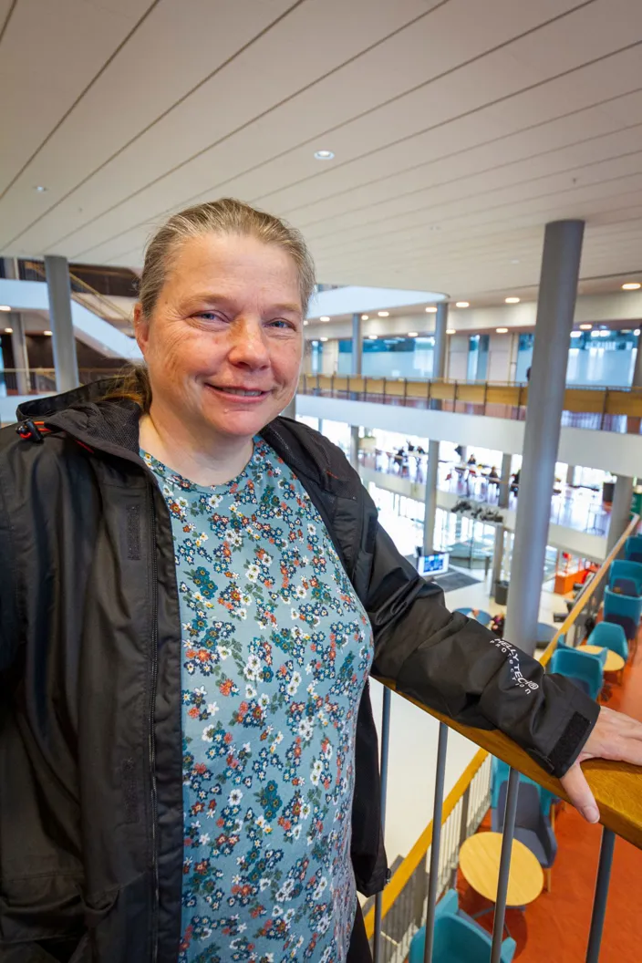 En kvinna står vid ett räcke i en stor, ljus byggnad med flera våningsplan och öppna ytor i bakgrunden. Hon bär en blommig tröja och en svart jacka och tittar mot kameran.