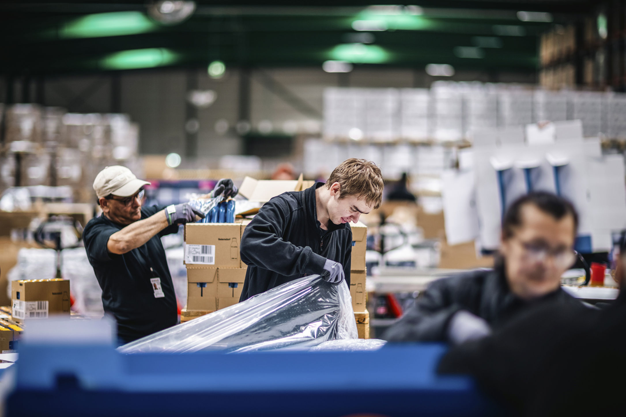 Flera medarbetare arbetar med packning och hantering av varor i en lager- och produktionsmiljö.