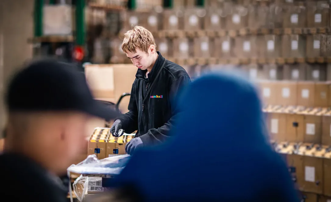 En medarbetare arbetar med paketering eller montering vid ett arbetsbord i en lager- eller produktionsmiljö. I bakgrunden syns staplade kartonger och lagerhyllor, medan andra personer syns oskarpt i förgrunden.