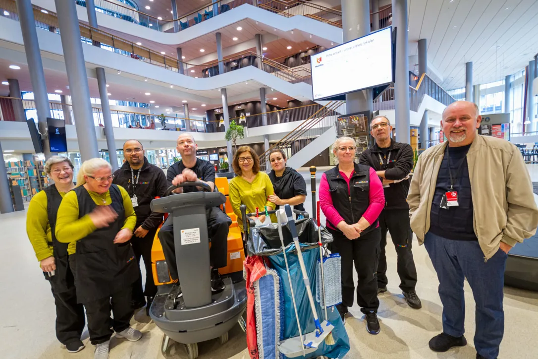 En grupp städmedarbetare står tillsammans i en stor, modern byggnad med flera våningsplan och öppna balkonger. I mitten syns en städmaskin och en städvagn med moppar och utrustning.