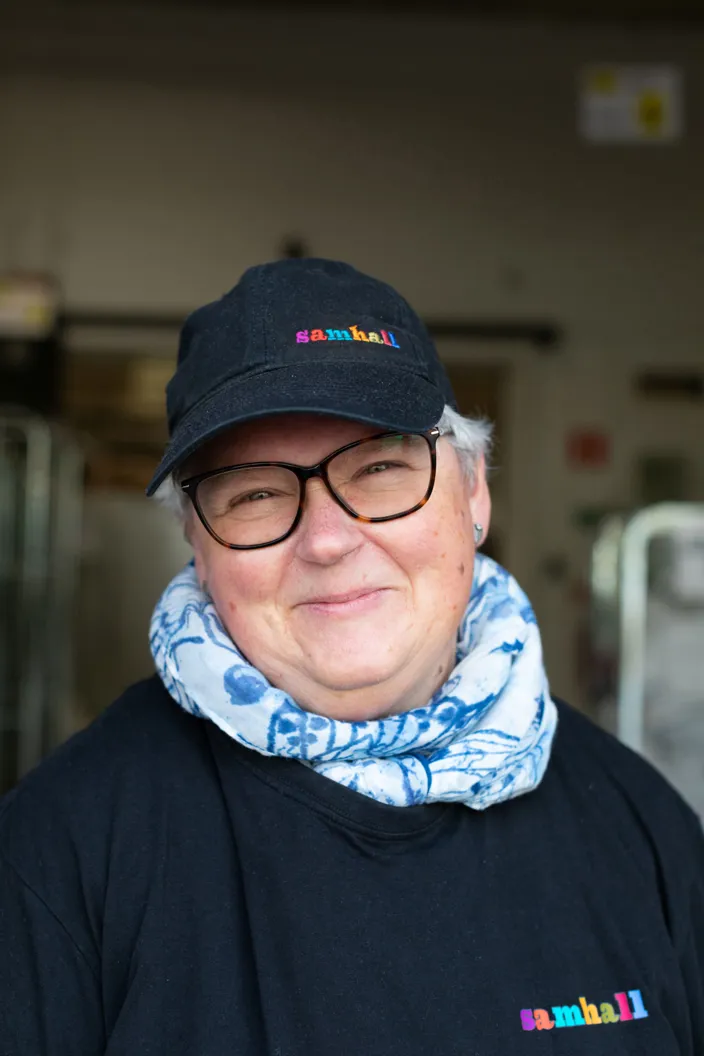 En kvinna med glasögon och en svart keps med Samhall-logotypen ler mot kameran. Personen bär en mörk tröja och en ljusblå mönstrad halsduk och står i en industrimiljö med utrustning i bakgrunden.