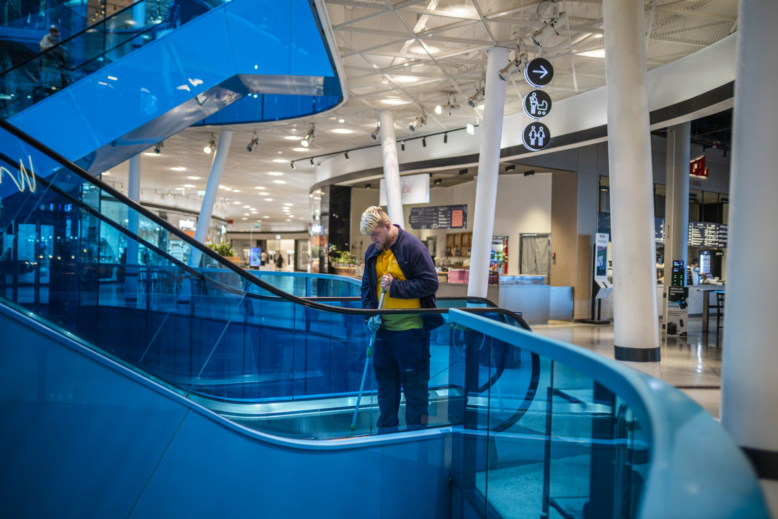 Person som arbetar med städning står på en rulltrappa i ett köpcentrum och moppar golvet. Rulltrappan har blå glasräcken och i bakgrunden syns butiker, skyltar och öppna ytor.