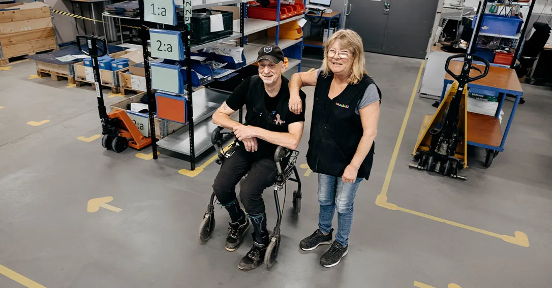 Bild på två medarbetare som står i ett lager. På golvet syns gula tejpmarkeringar. Medarbetarna är klädda i svart och tittar in i kameran. En står och en sitter i en rullstol. Bakom personerna står det hyllor med prylar.