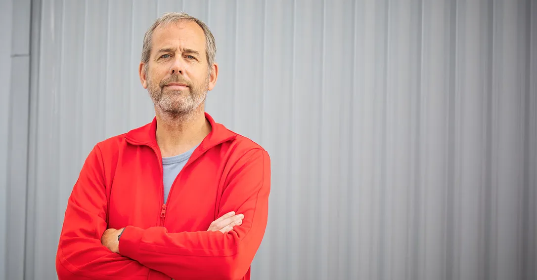 Jörgen Lorentzon med ljusbrunt hår och gråspräckligt skägg samt blå ögon står med armarna i kors och tittar självsäkert in i kameran framför en grå plåtvägg