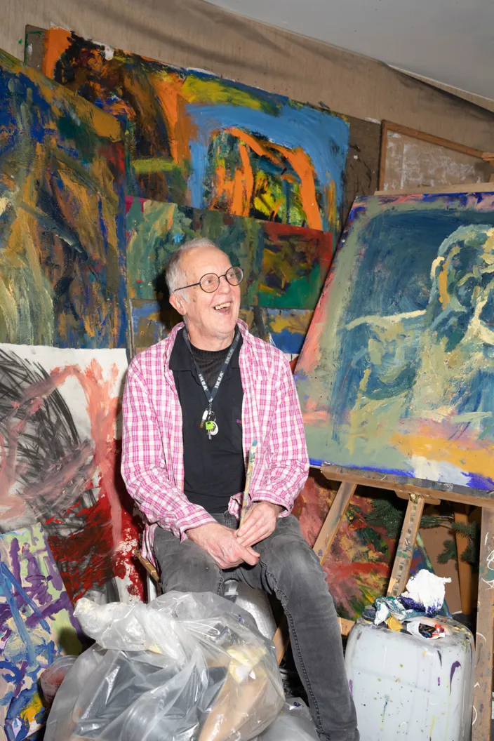 Thomas sitter på en stol i en konstnärsateljé, omgiven av stora färgstarka målningar lutade mot väggarna. Han bär en rosa rutig skjorta över en mörk tröja och ler medan han tittar åt sidan. I förgrunden syns plastpåsar och målarutrustning.