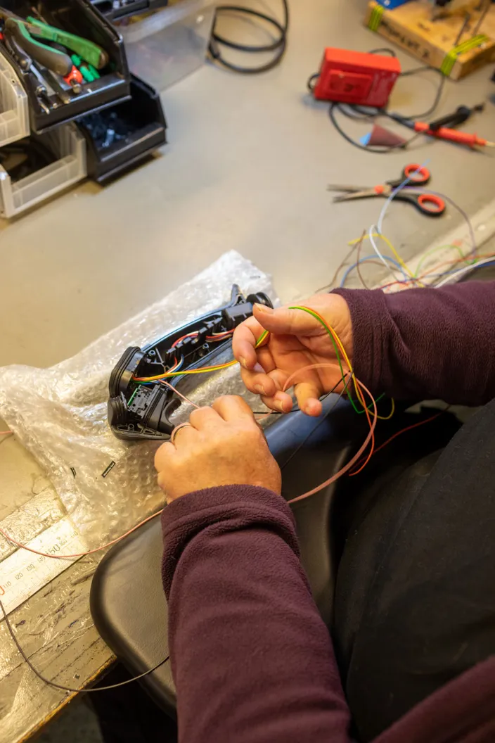 Närbild av ett par händer som arbetar med färgade elkablar inuti en öppnad plastkomponent på ett arbetsbord. Komponenten ligger på bubbelplast, och runt omkring syns verktyg som sax, kablar och förvaringslådor med delar.