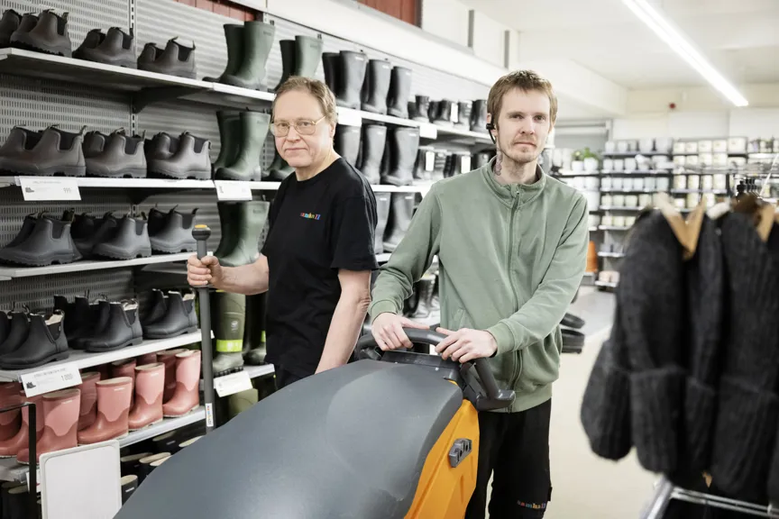 Två medarbetare arbetar i en butik med hyllor fyllda av stövlar och skor. En person kör en städmaskin medan den andra står bredvid med ett redskap i handen.