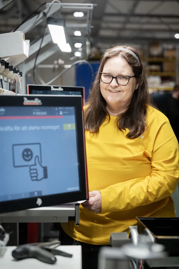 En medarbetare står vid en monteringsstation i en industrimiljö. På en skärm bredvid visas instruktioner för arbetsmomentet.