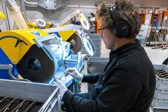 En medarbetare arbetar vid en maskin för bearbetning av glasögonbågar i en industrimiljö. Personen använder hörselskydd och håller i ett par glasögon.