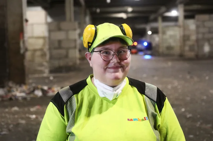 Porträttbild på Jennifer som ler och tittar in i kameran. Hon bär arbetskläder i reflex och har rölurar uppsatta på huvudet. 