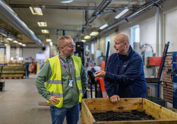 Två kollegor samtalar i en industrimiljö vid en arbetsstation med material i en trälåda.