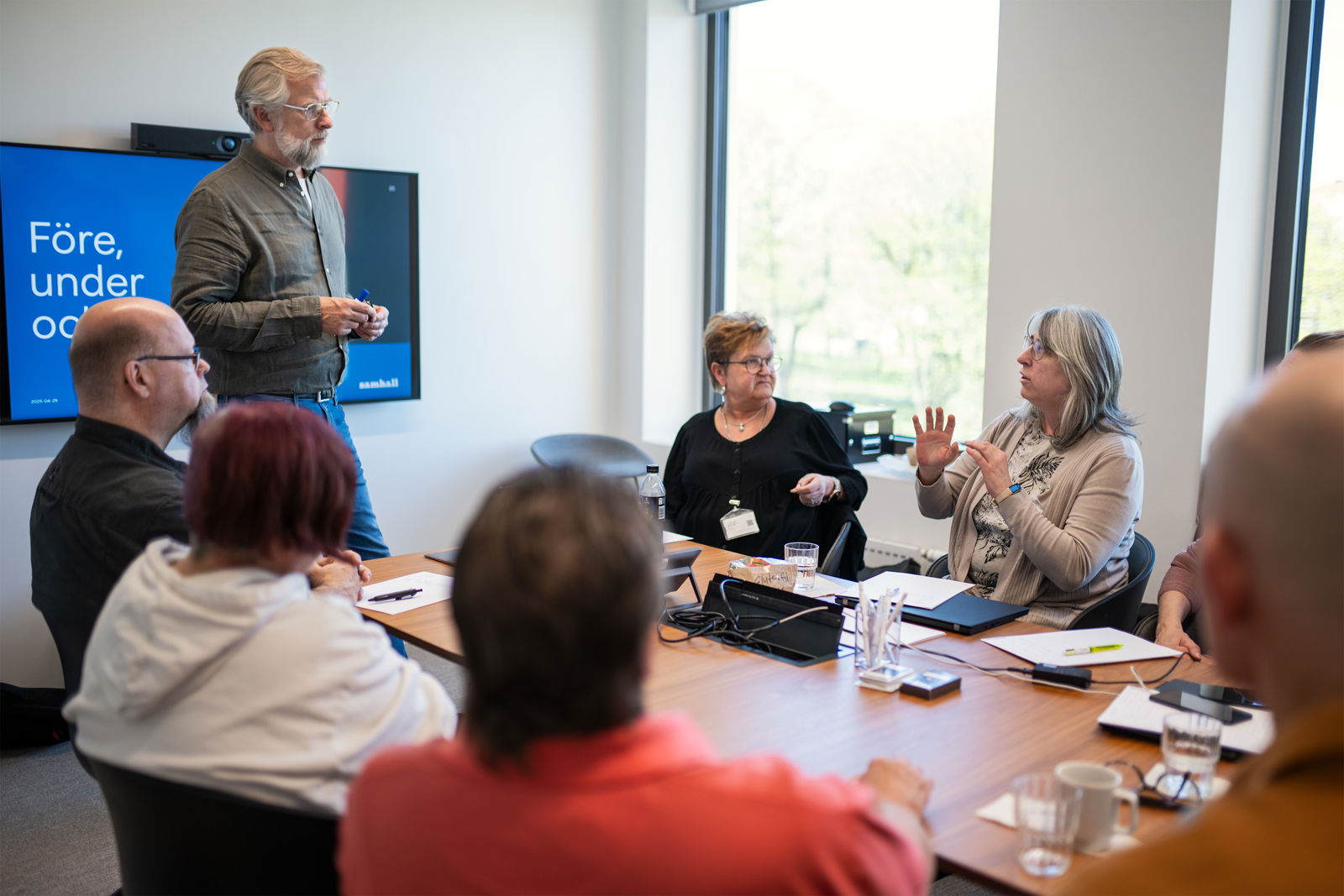 En grupp samhallsmedarbetare sitter runt ett bord i ett mötesrum medan en person står och presenterar vid en skärm.