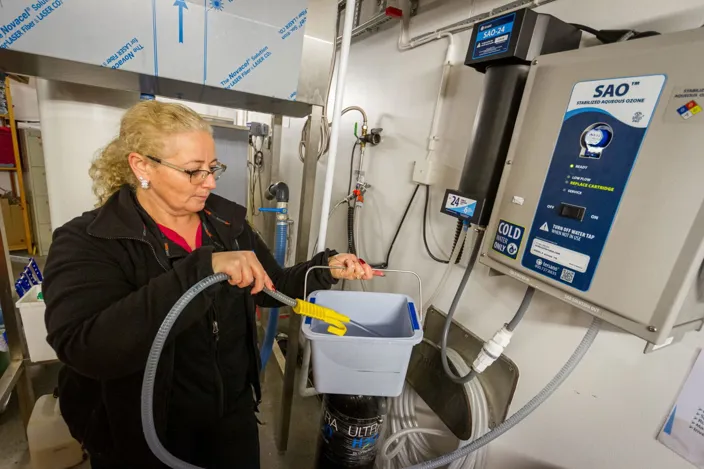 En kvinna fyller en hink med vatten via en slang kopplad till en väggmonterad enhet märkt ”SAO – Stabilized Aqueous Ozone” i ett teknikutrymme.