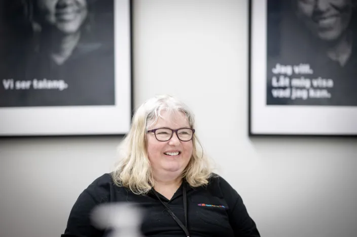 Kvinna med glasögon, ljust hår och svart Samhalltröja. Hon ler och tittar åt sidan. I bakgrunden syns två tavlor suddigt.
