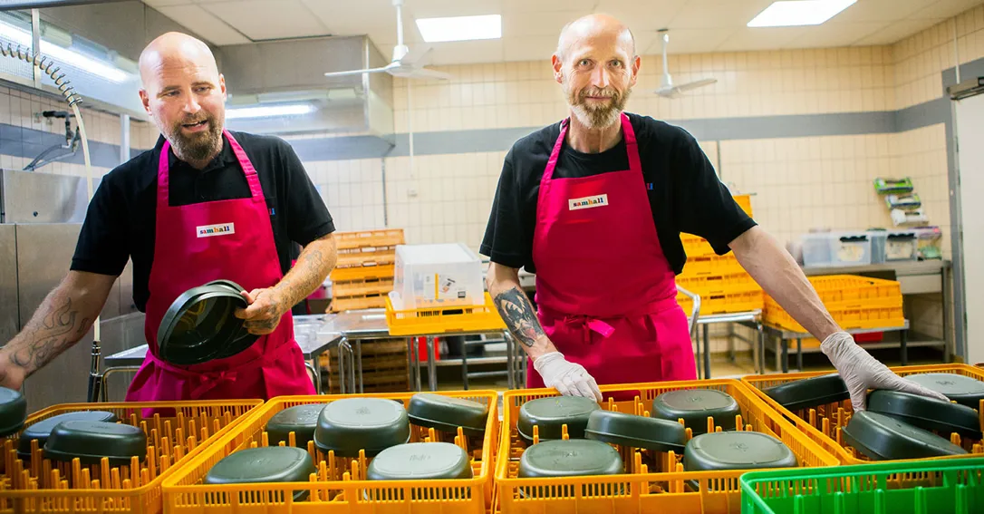 Bild på två personer med lila förkläden som fixar med disk i ett kök.