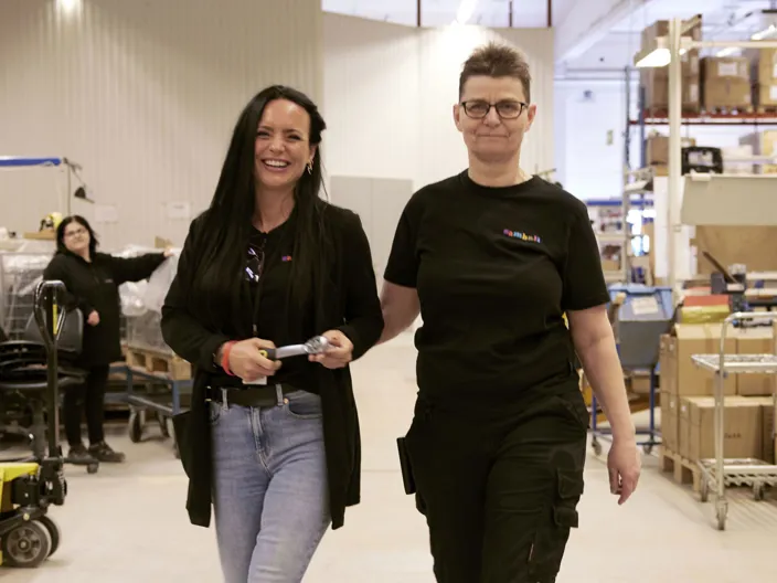 Bild på tre kvinnor i Samhallkläder. En i bakgrunden, två i förgrunden. De går i en lagerlokal.