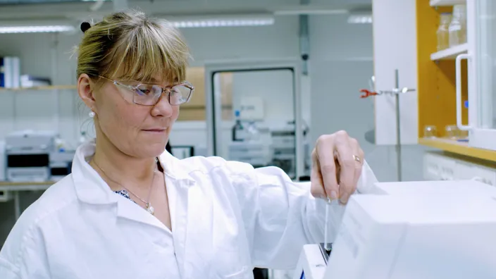 En kvinna i vit laboratorierock och skyddsglasögon arbetar i ett laboratorium. Hon håller i ett provrör som placeras i en laboratoriemaskin, med hyllor och utrustning i bakgrunden.