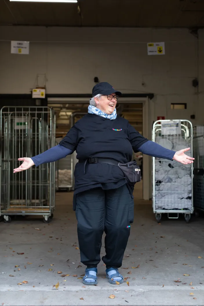 En person i arbetskläder med Samhall-logotypen står med armarna utsträckta och ler. Personen bär keps, glasögon och halsduk och står i en industrimiljö framför stora metallvagnar fyllda med textilier.