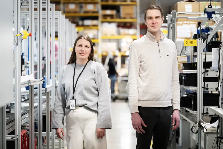 Två personer står i en produktionsmiljö mellan monteringsstationer och hyllsystem. I bakgrunden syns arbetsutrustning, komponenter och lagerhyllor.