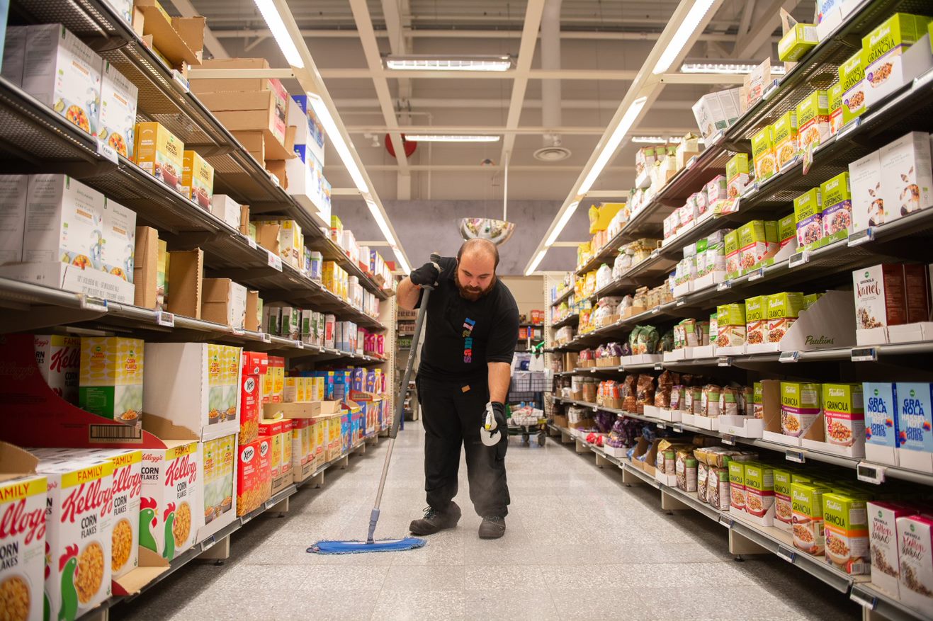 En person städar golvet med en mopp i en livsmedelsbutik, omgiven av hyllor fyllda med frukostflingor och andra matvaror.