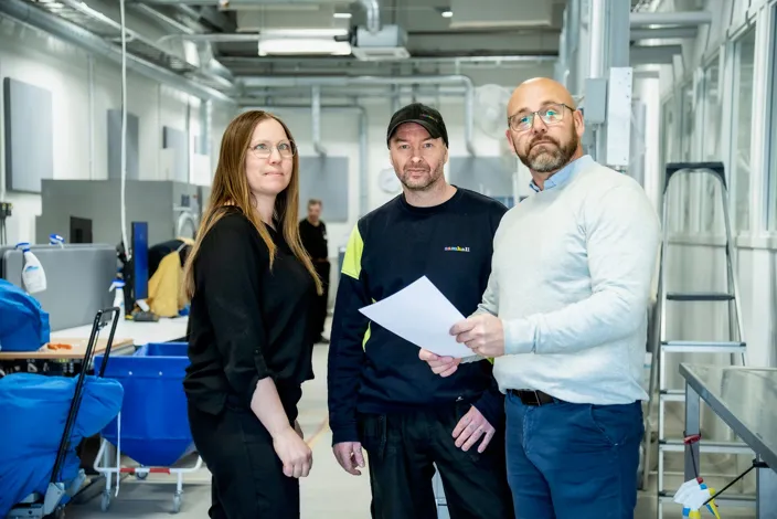 Tre personer står i en ljus industrimiljö. En man håller ett papper medan de andra två står bredvid och tittar mot kameran. I bakgrunden syns arbetsstationer, utrustning och ventilationsrör i taket.