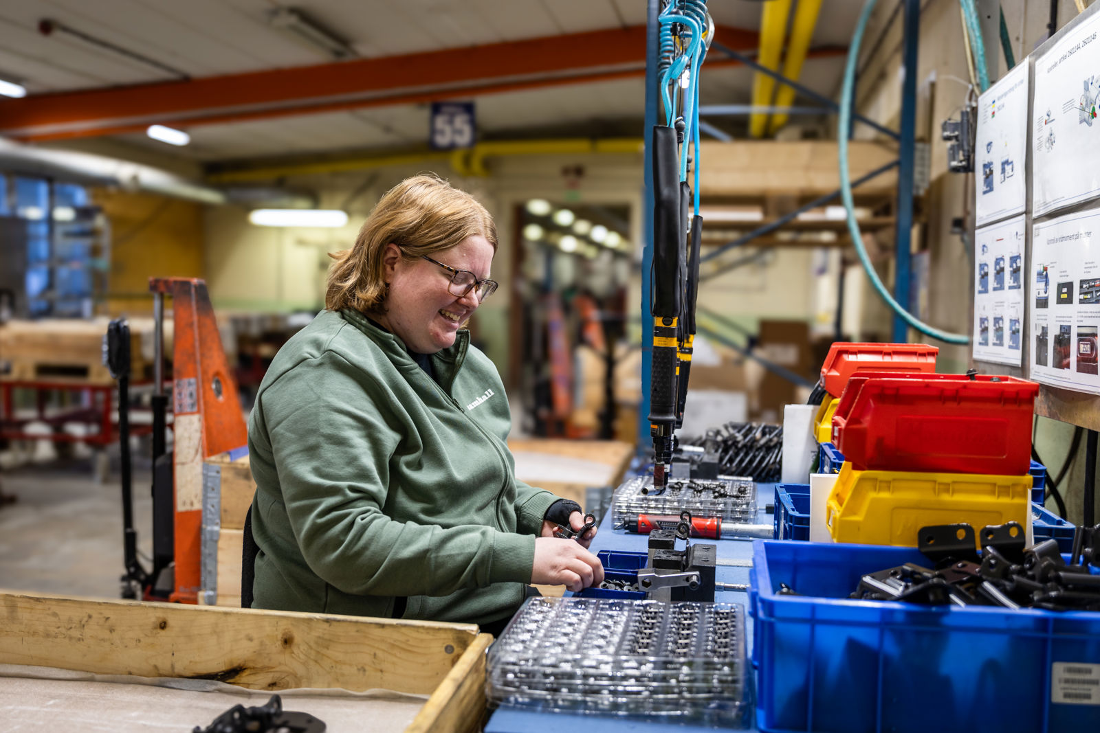 Samhalls medarbetare Marie sitter vid en station och monterar gångjärn till lastbilar på Beslag och Metall i Vetlanda. Hon har ljust hår, glasögon och en grön Samhall-tröja. Hon ler.