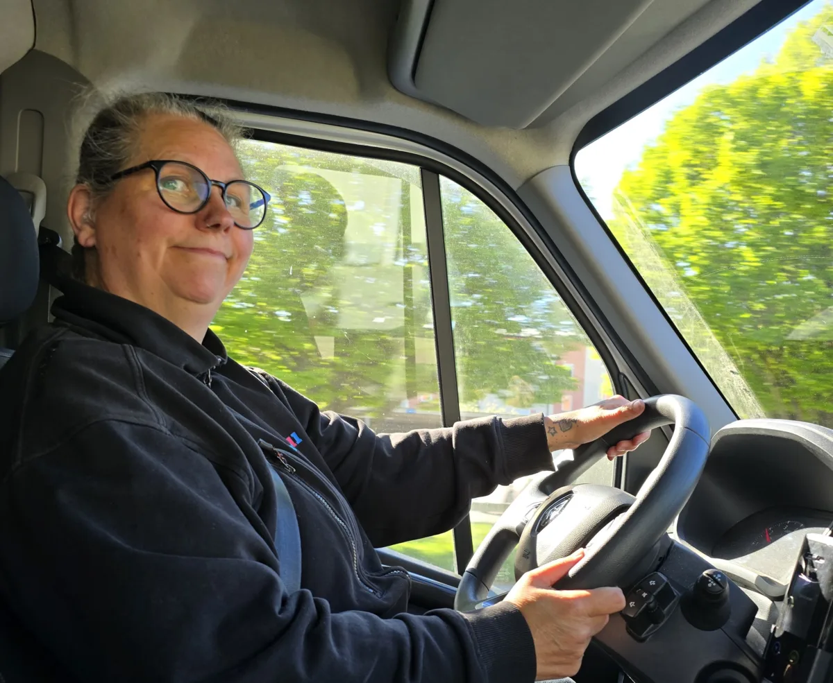 Kvinna med glasögon sitter i förarsätet och kör en skåpbil. Hon håller i ratten och tittar framåt. Utanför bilfönstret syns gröna träd i dagsljus.