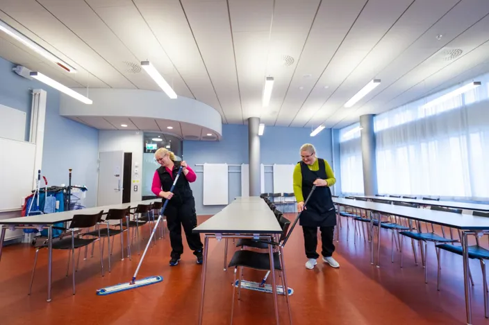 Två städmedarbetare moppar golvet i en ljus samlingssal med uppställda bord och stolar i rader.