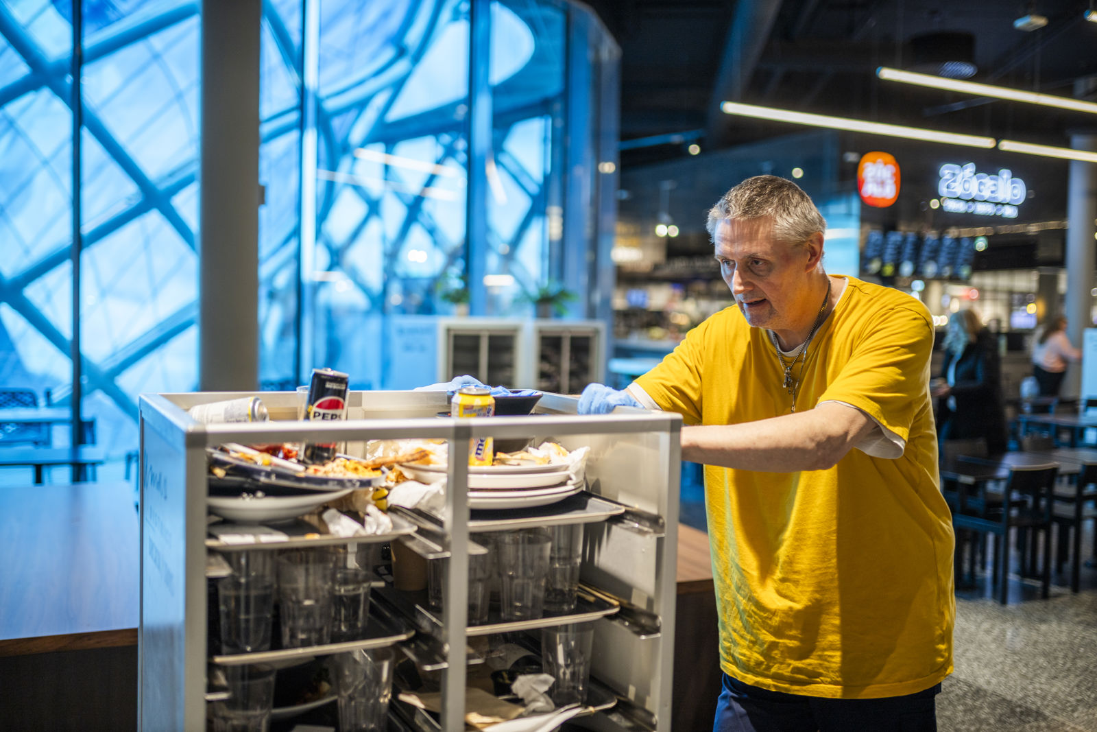 Samhallsmedarbetare i gul tröja kör en vagn med disk i en restaurangmiljö.