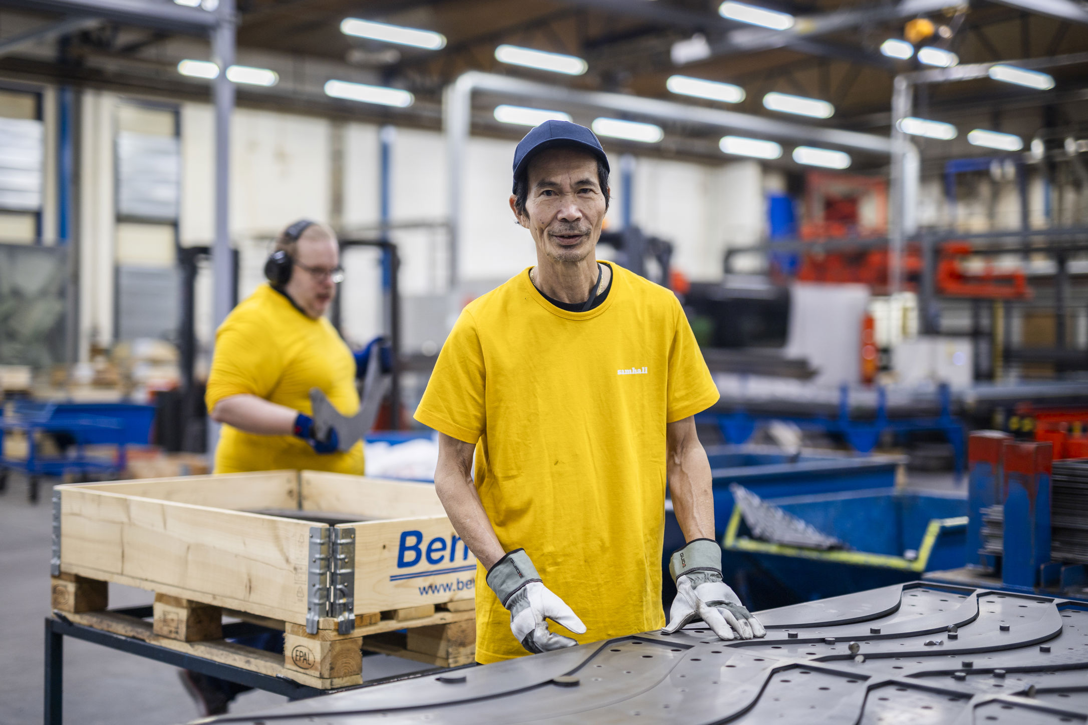 Samhallsmedarbetare arbetar vid en maskin i en industrimiljö, med en kollega i bakgrunden.