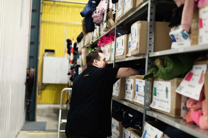 Jimmy står emd ryggen vänd mot karmen och rotar i en låd i ansiktshöjd på en lagerhylla. 