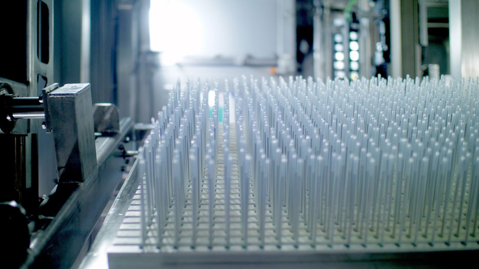 Rader av tunna, genomskinliga plastdetaljer står tätt uppradade på en bricka i en industriell miljö. Maskindelar och starkt ljus syns suddigt i bakgrunden.
