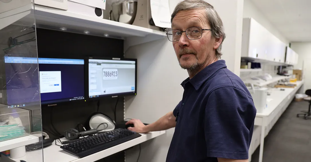 En man med glasögon tittar in i kameran. Han står vid två datorskärmar och arbetar.