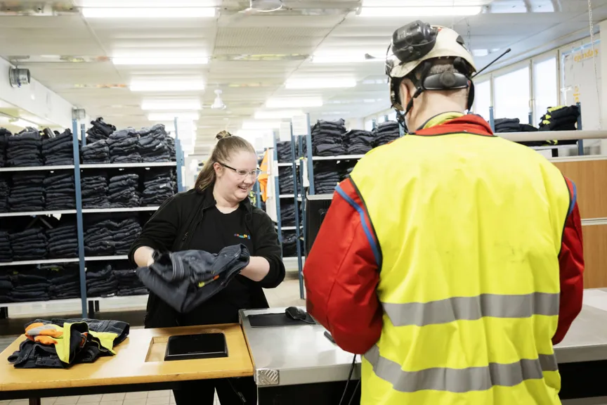 En medarbetare lämnar ut arbetskläder till en person i varselkläder och hjälm. I bakgrunden syns hyllor med vikta arbetsplagg.