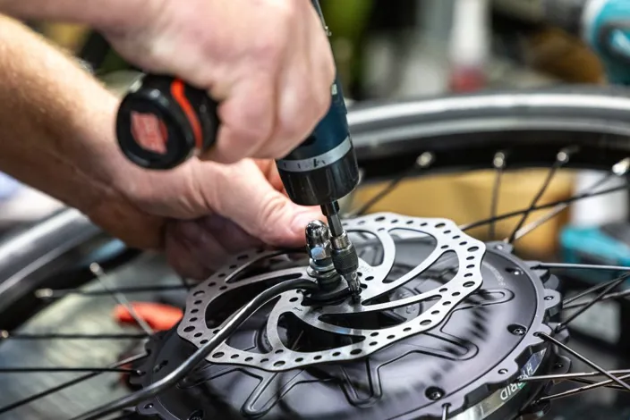 Närbild på en samhallsmedarbetare som använder en skruvdragare för att montera en bromsskiva på ett cykelhjul.