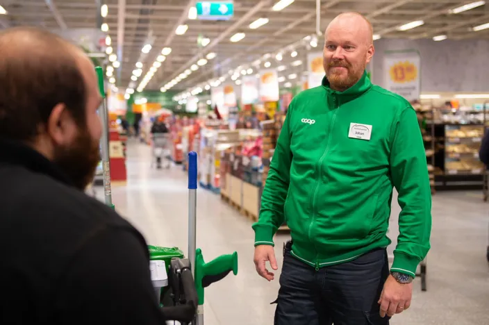 En butikschef i grön Coop-jacka med namnskylt står i en livsmedelsbutik och samtalar med en städmedarbetare bredvid en städvagn.