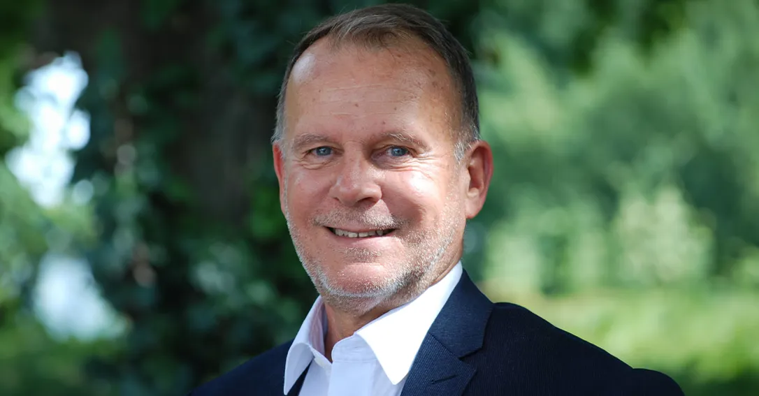 Bild på en man med mörk kavaj och vit skjorta. Han ler och tittar in i kameran. Bakom honom syns grönskande natur suddigt.