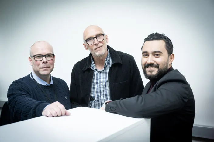 tre män står lutade mot en vit möbel och tittar in i kameran. De ser vänliga ut. Två bär glasögon. 