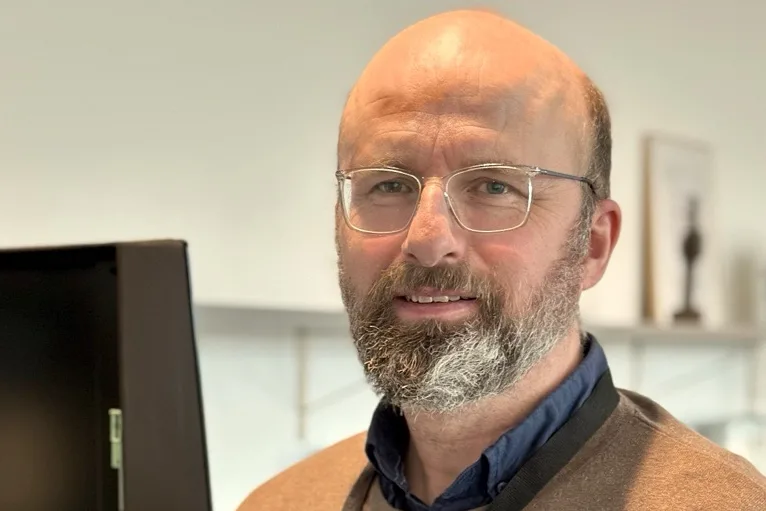 Porträtt av en vuxen man med glasögon och skägg som står inomhus vid en datorskärm. Han bär skjorta och tröja och tittar mot kameran. I bakgrunden syns en ljus kontorsmiljö med hyllor.