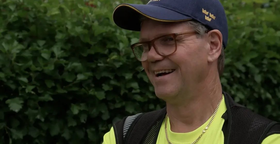 En man i keps och glasögon ler utomhus med grönska i bakgrunden.