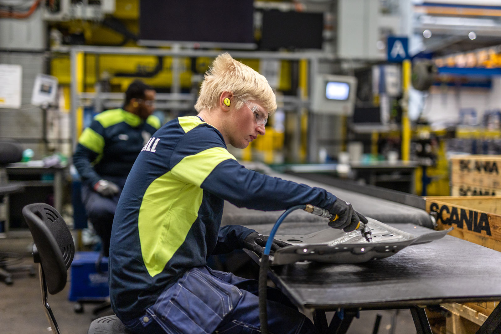 Person arbetar med ett metallföremål vid ett arbetsbord i industrimiljö.