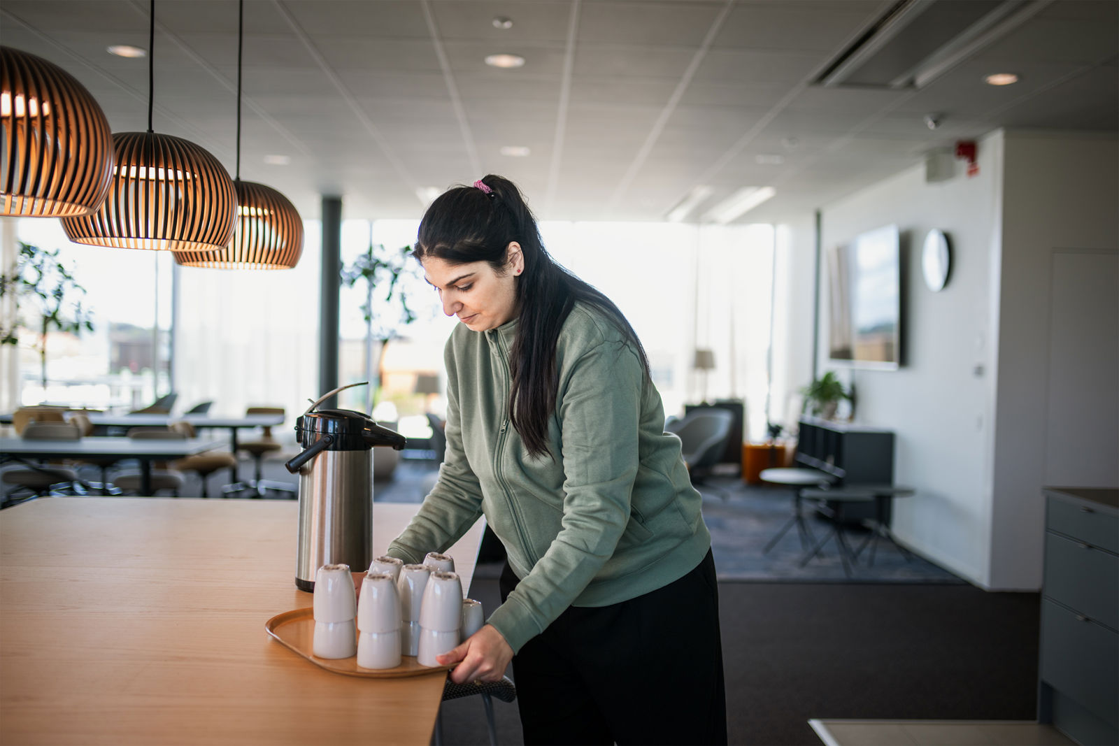 Kvinna ställer fram koppar och termos på ett bord i en kontorsmiljö.