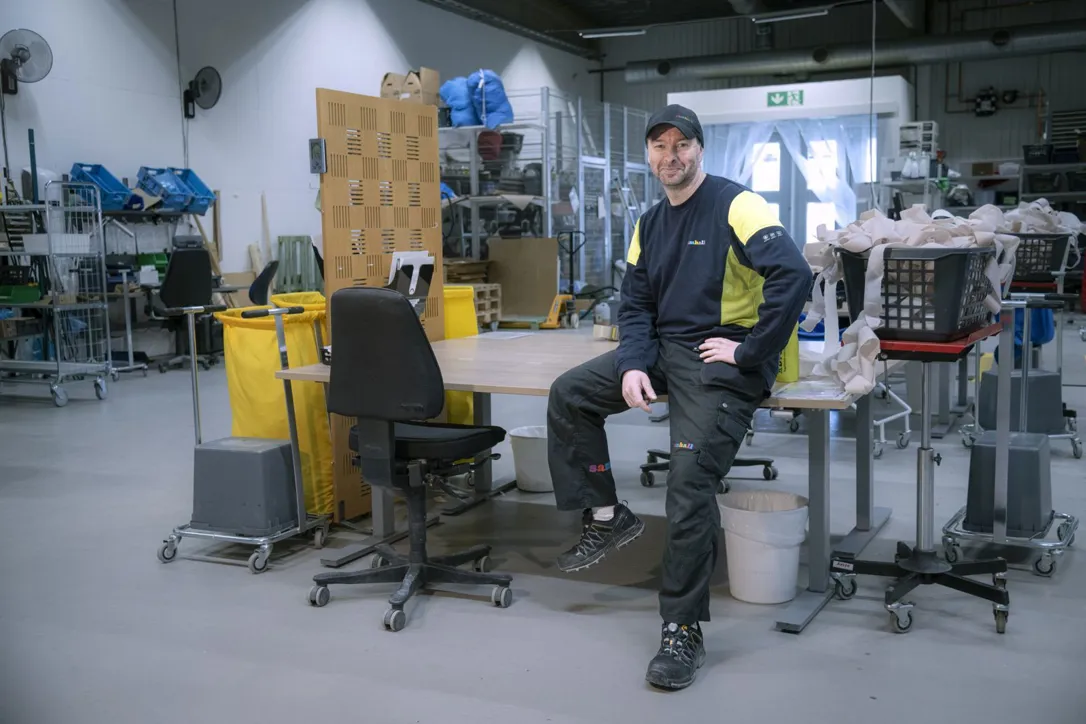 En Samhallmedarbetare sitter vid en arbetsstation i en verkstadsmiljö omgiven av utrustning och material.