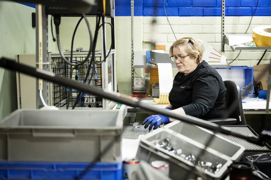 En medarbetare sitter vid en arbetsstation i en industrimiljö och arbetar med mindre metalldelar. På arbetsbordet finns verktyg, plastlådor och komponenter.