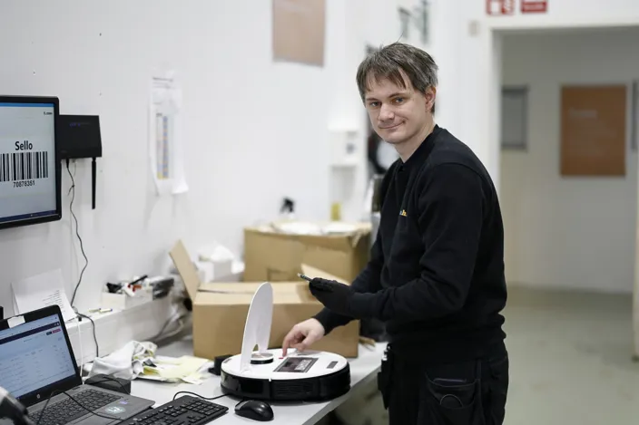 Medarbetaren Alexander står vid ett bord och mekar med en robotdammsugare. Han tittar in i kameran. 