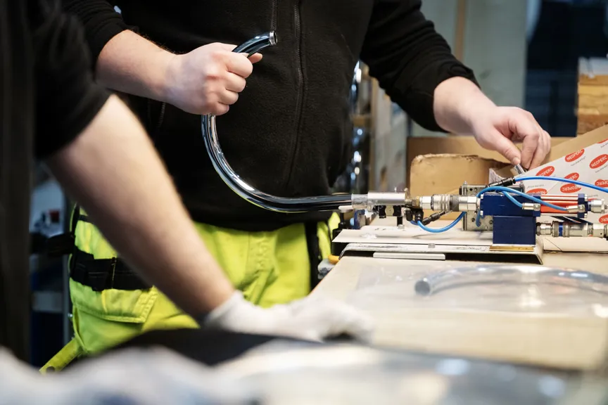 Två personer arbetar med montering av metalldelar vid en arbetsbänk. En person håller i en böjd metallkomponent kopplad till en maskin.