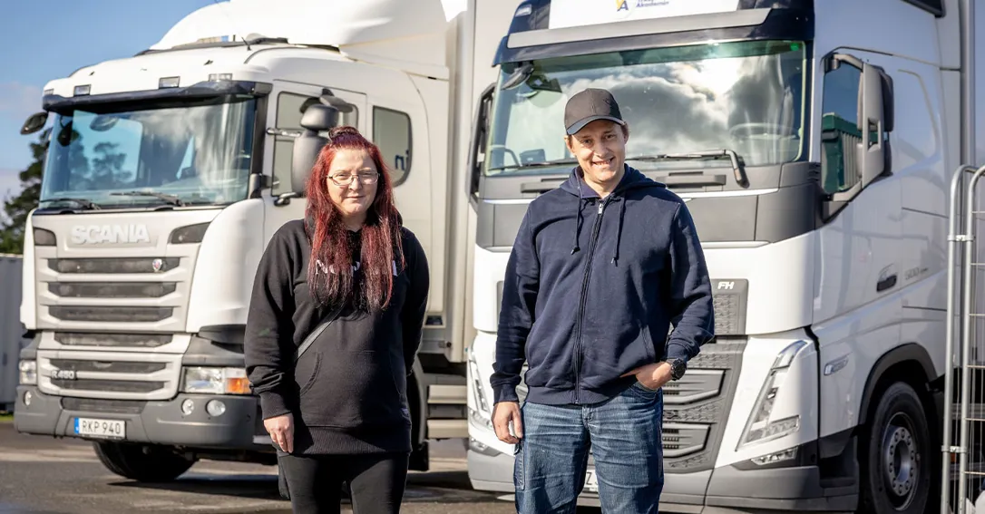 Två personer står framför lastbilar på en uppställningsplats och tittar mot kameran.